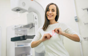 Woman holding breast cancer ribbon in front of mammogram machine.